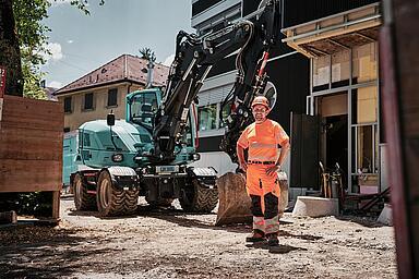 Un opérateur vêtu d'une tenue de travail haute visibilité se tient à côté d'une pelle sur pneus Wacker Neuson EW100 sur un chantier urbain.