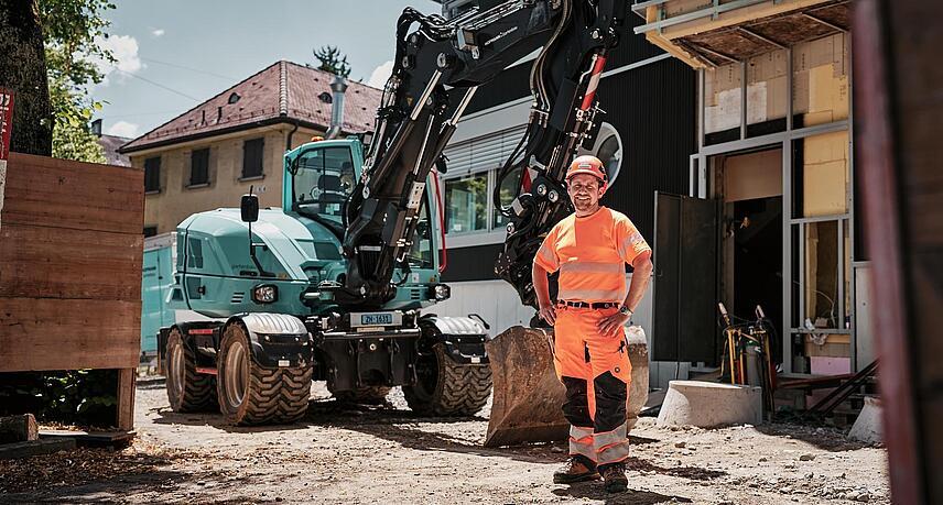 Een machinist in goed zichtbare werkkleding staat naast een Wacker Neuson EW100-graafmachine op wielen op een bouwplaats in de stad.