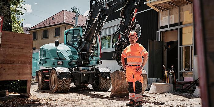 Een machinist in goed zichtbare werkkleding staat naast een Wacker Neuson EW100-graafmachine op wielen op een bouwplaats in de stad.
