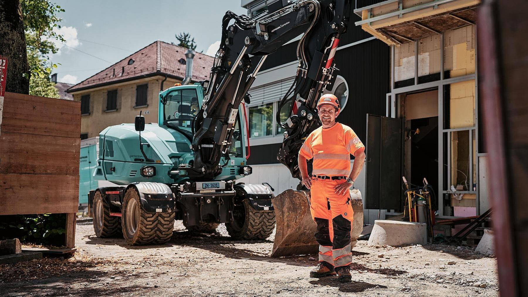 Een machinist in goed zichtbare werkkleding staat naast een Wacker Neuson EW100-graafmachine op wielen op een bouwplaats in de stad.