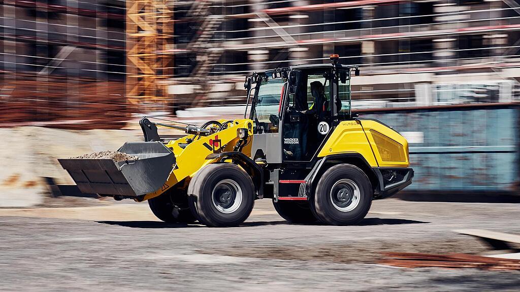 Wacker Neuson WL950p wheel loader transporting material across a construction site in front of a building under construction.