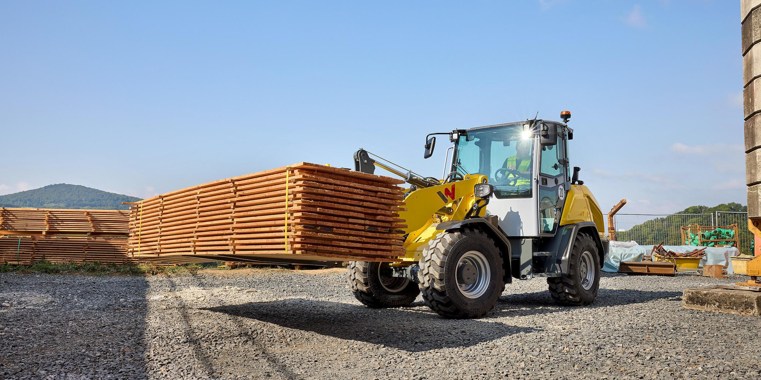 Wacker Neuson WL950 wheel loader transporting a stack of timber boards with pallet forks in a construction materials yard.