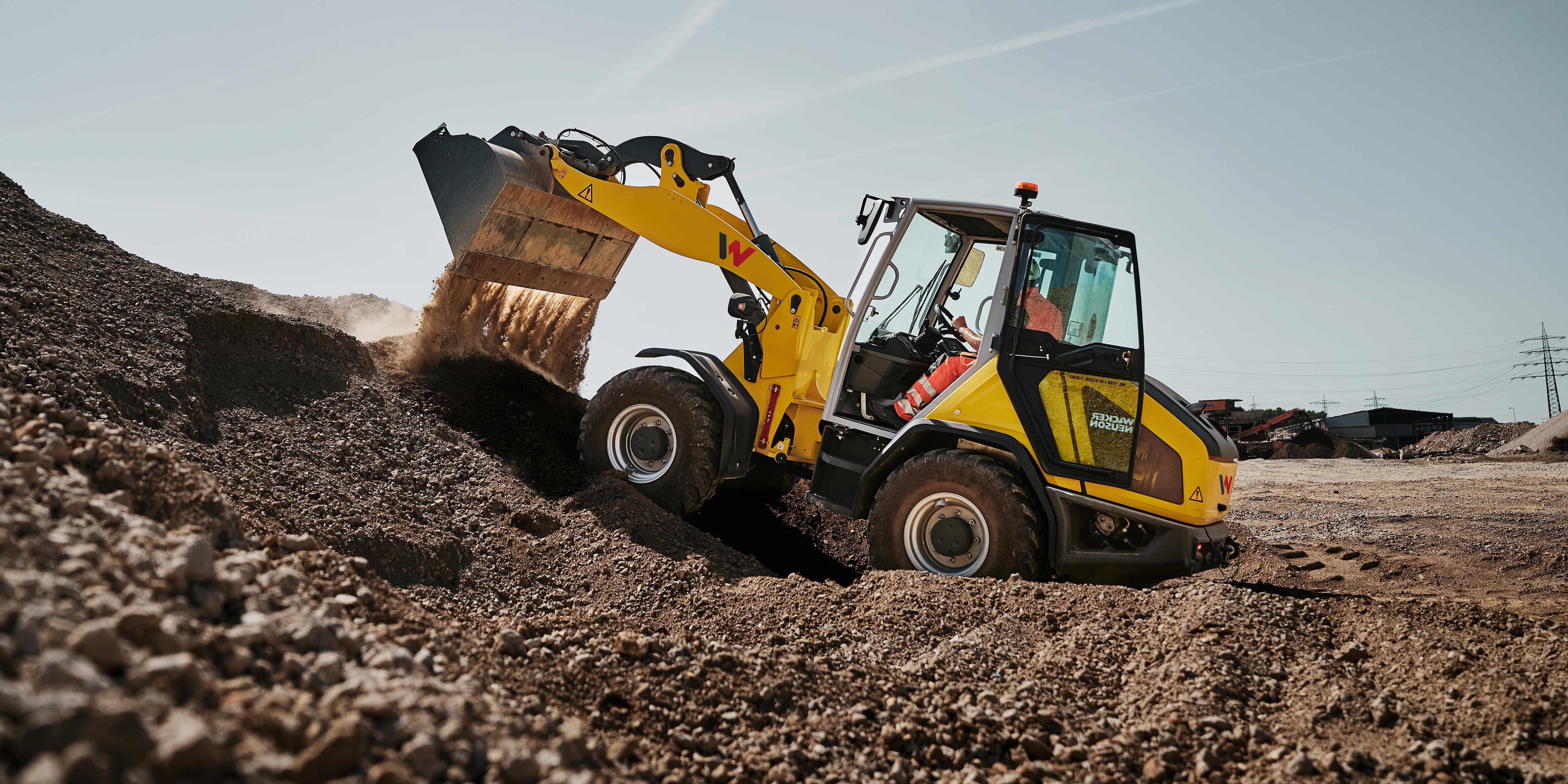 Wacker Neuson WL1150 wheel loader loading and moving aggregate material at a quarry site, with the bucket raised and material flowing from the bucket.