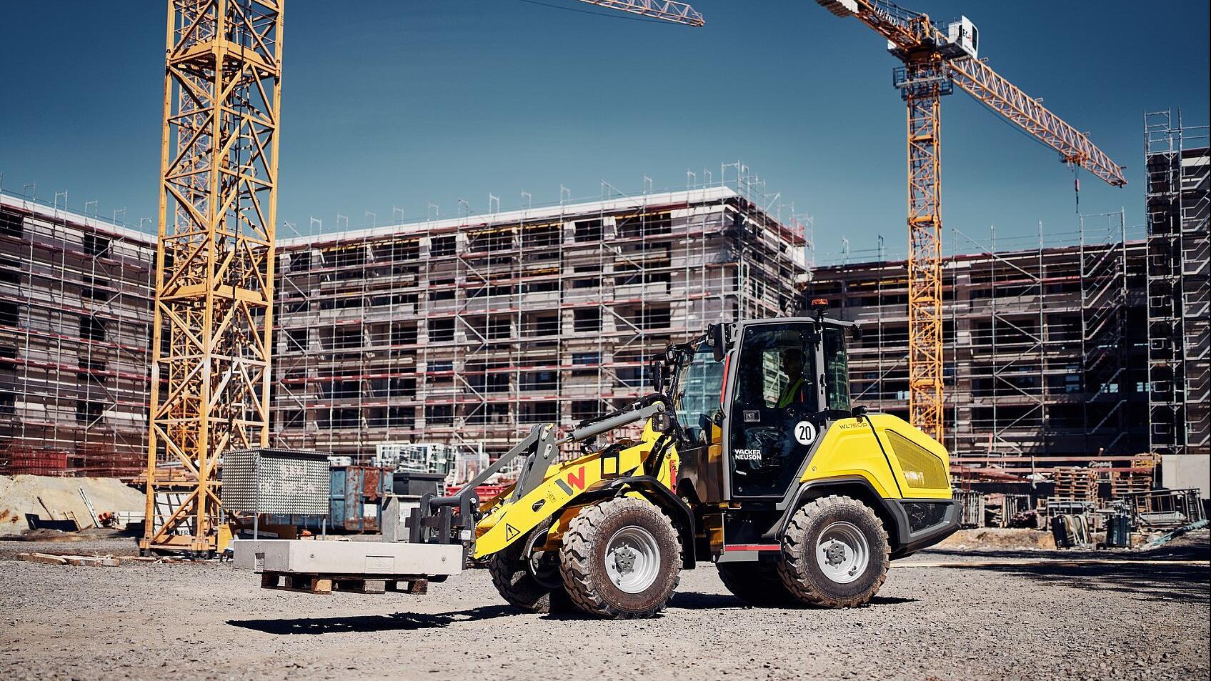 Une chargeuse sur pneus Wacker Neuson WL750p transportant des éléments en béton sur un chantier en activité, avec en arrière-plan des échafaudages et des grues à tour.