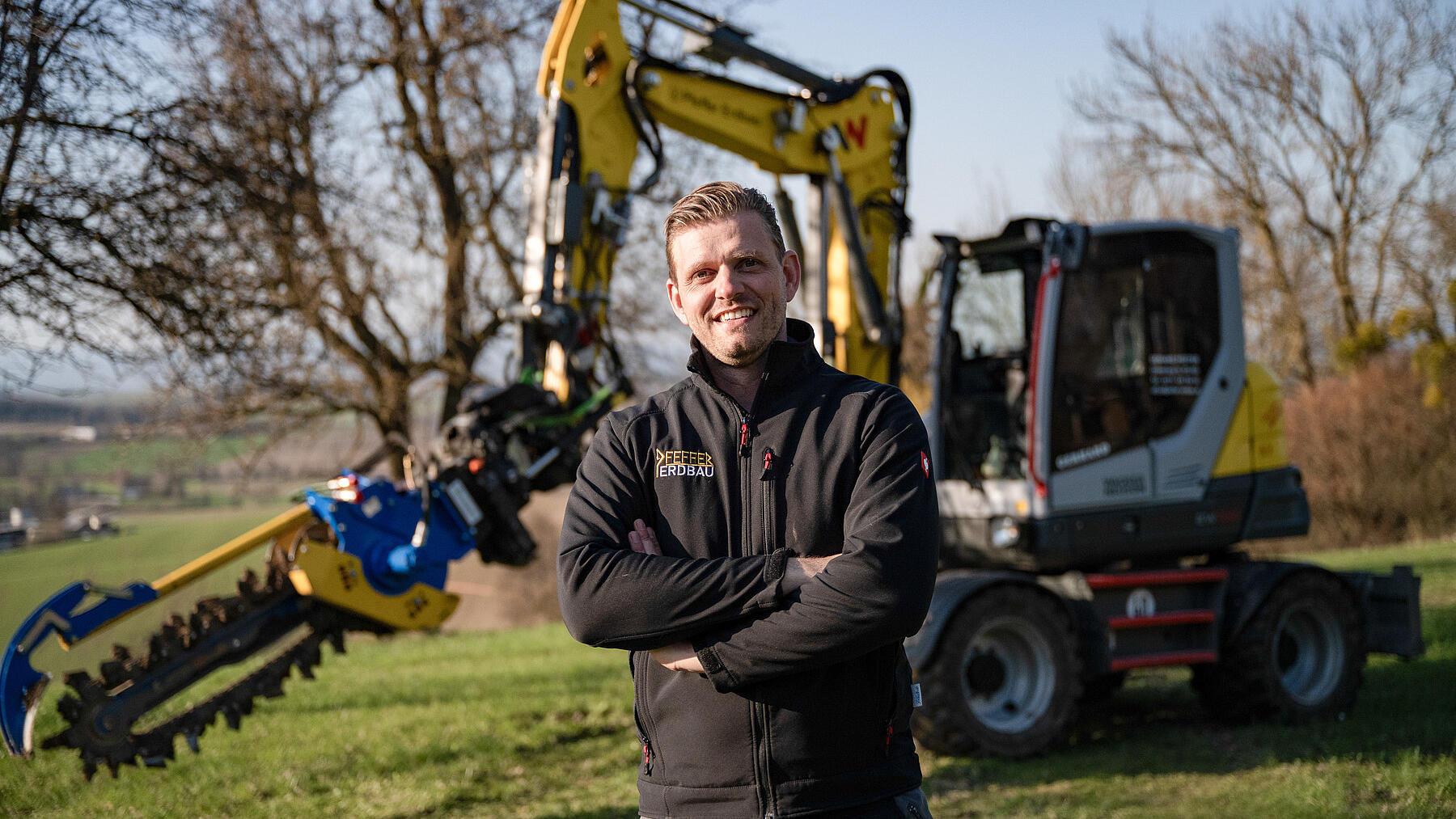 Een aannemer staat voor een Wacker Neuson EW100-graafmachine op wielen, uitgerust met een sleufgraafinrichting, op een werkterrein op het platteland.