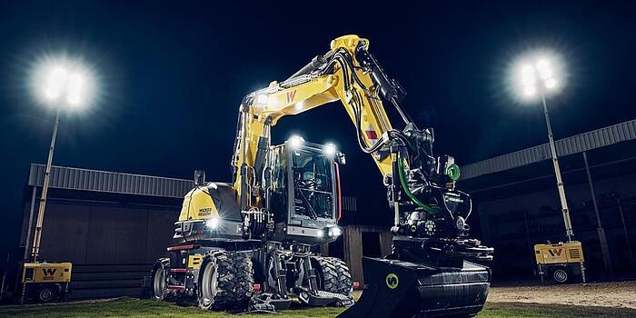 Pelle sur pneus Wacker Neuson EW135 équipée d'un rotateur inclinable et d'un godet de nivellement, éclairée par des projecteurs dans un environnement de travail nocturne.c