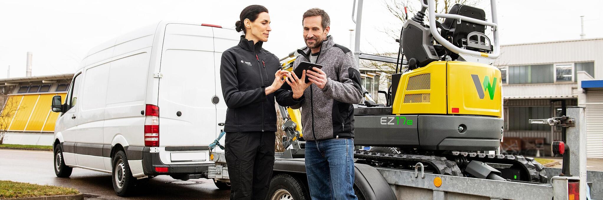 Des articles de merchandising Wacker Neuson sont posés sur une table au premier plan, collaborateur Wacker Neuson en arrière-plan.