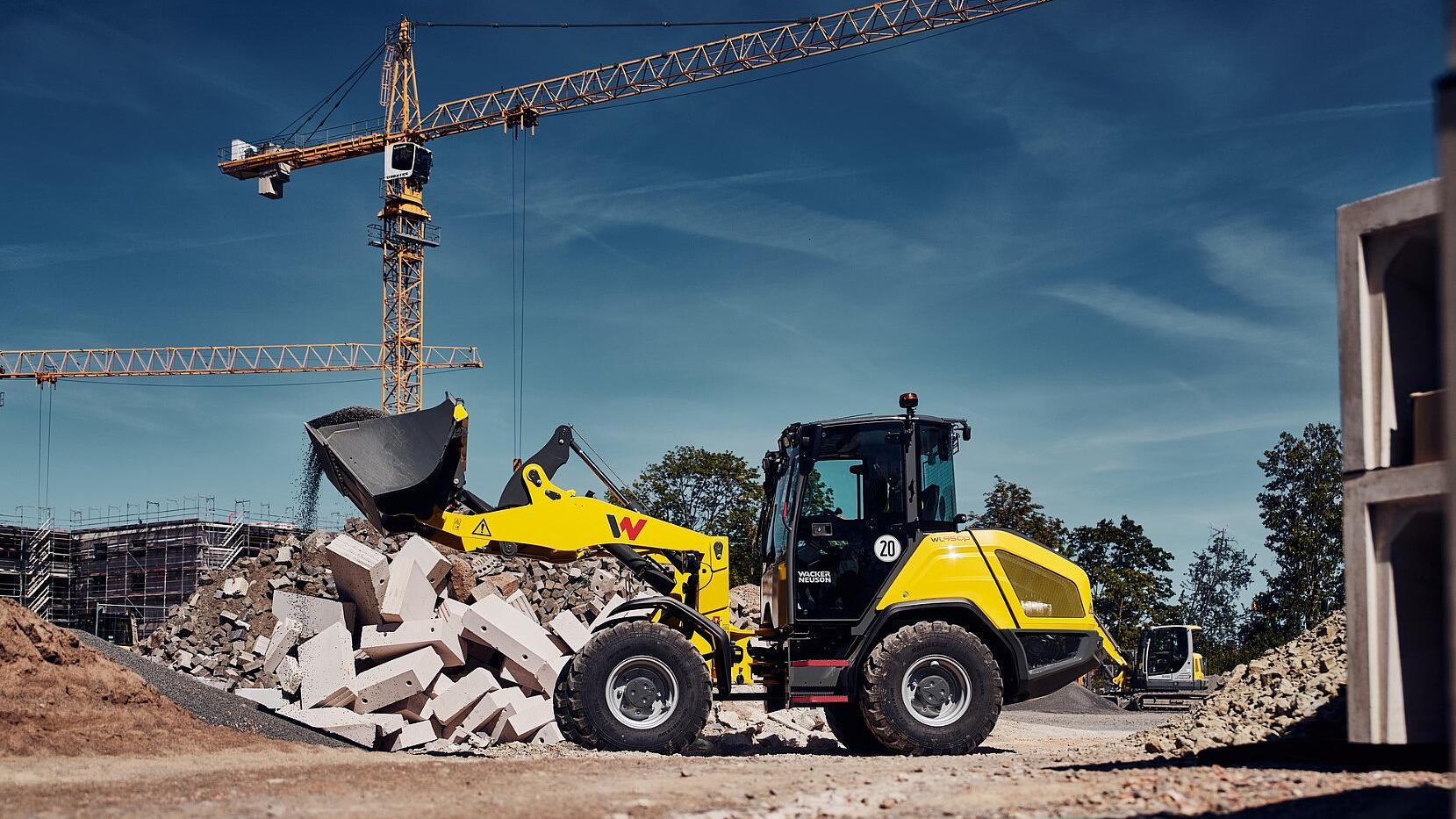 Une chargeuse sur pneus Wacker Neuson WL950p décharge des gravats de son godet sur un tas de débris de démolition sur un chantier de construction, avec des grues à tour en arrière-plan.