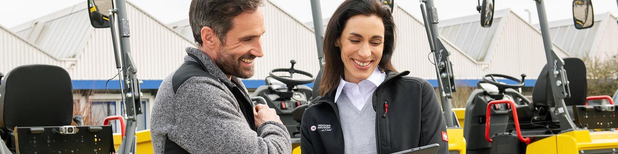 Une collaboratrice Wacker Neuson souriante et portant une tablette conseille un client souriant devant des machines de construction Wacker Neuson.